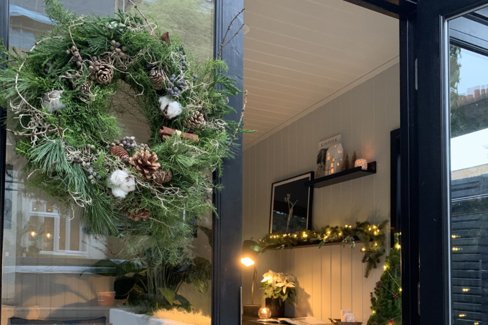A Christmas wreath hangs on the glass door of a painted garden studio. Inside, there are fairy lights and candles and you can see the styling is Christmassy