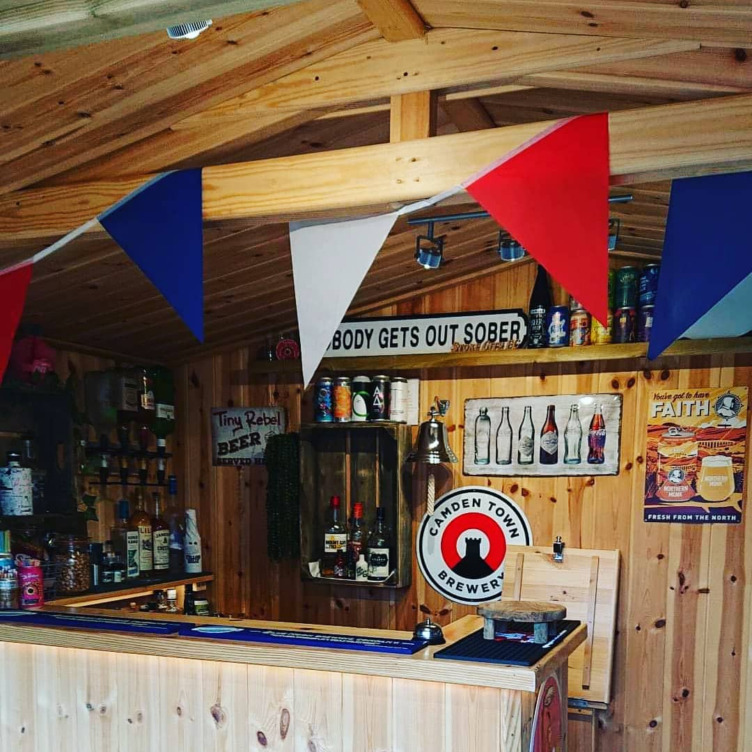 The interior of a wooden garden building that has red, white and blue bunting strung up. There is a wooden bar stocked with beers and spirits. On the wall there is a sign that says "nobody gets out sober".