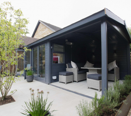 A garden building with an open sided area with comfortable lounge seating, surrounded by tidy planting