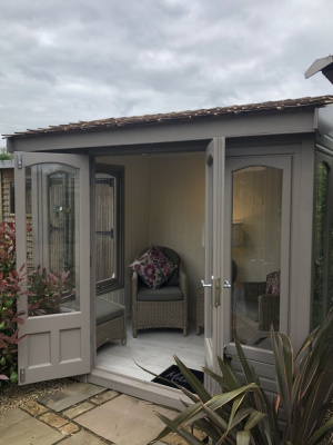 A small light grey rectangular summerhouse. The windows are full length and the double doors are open to show the cosy interior.