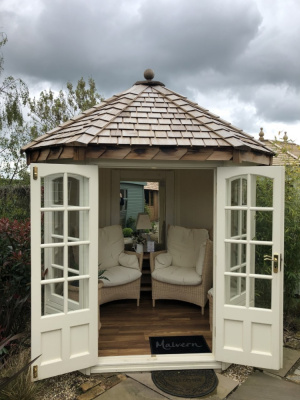A small, white square summerhouse with Georgian windows and double doors. The roof is hipped and made of cedar shingles. The double doors are open to reveal a cosy interior