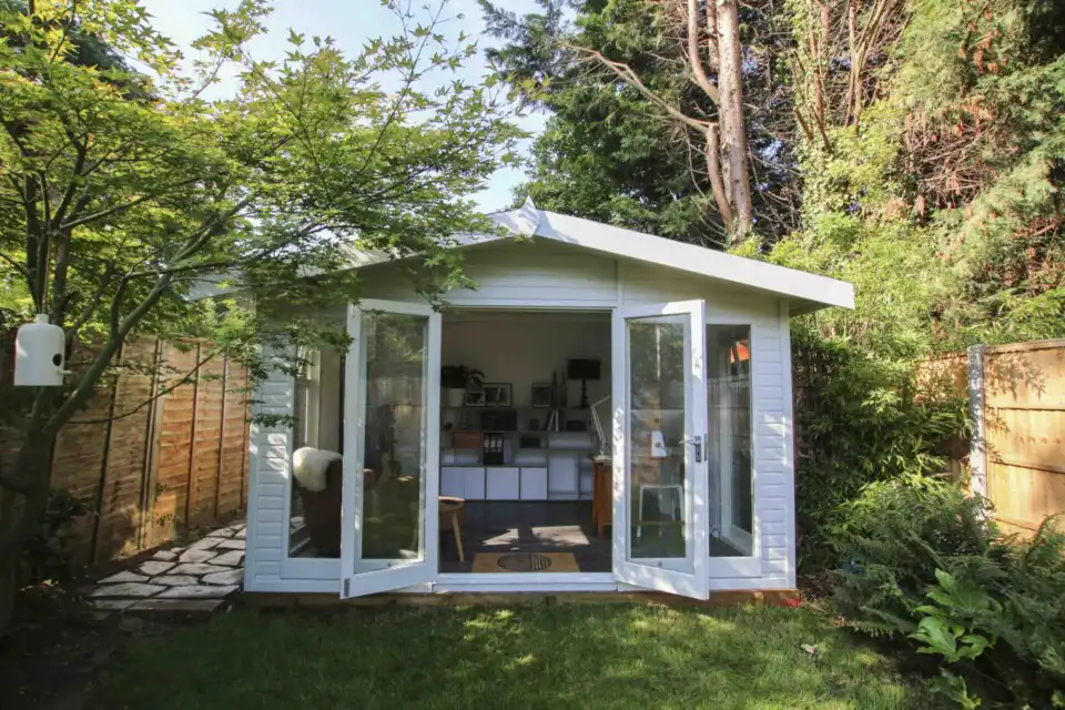 The image depicts a compact Studio Apex garden room by Malvern Garden Buildings, in a garden with a timber boundary fences. This customer used their garden building as a music room.