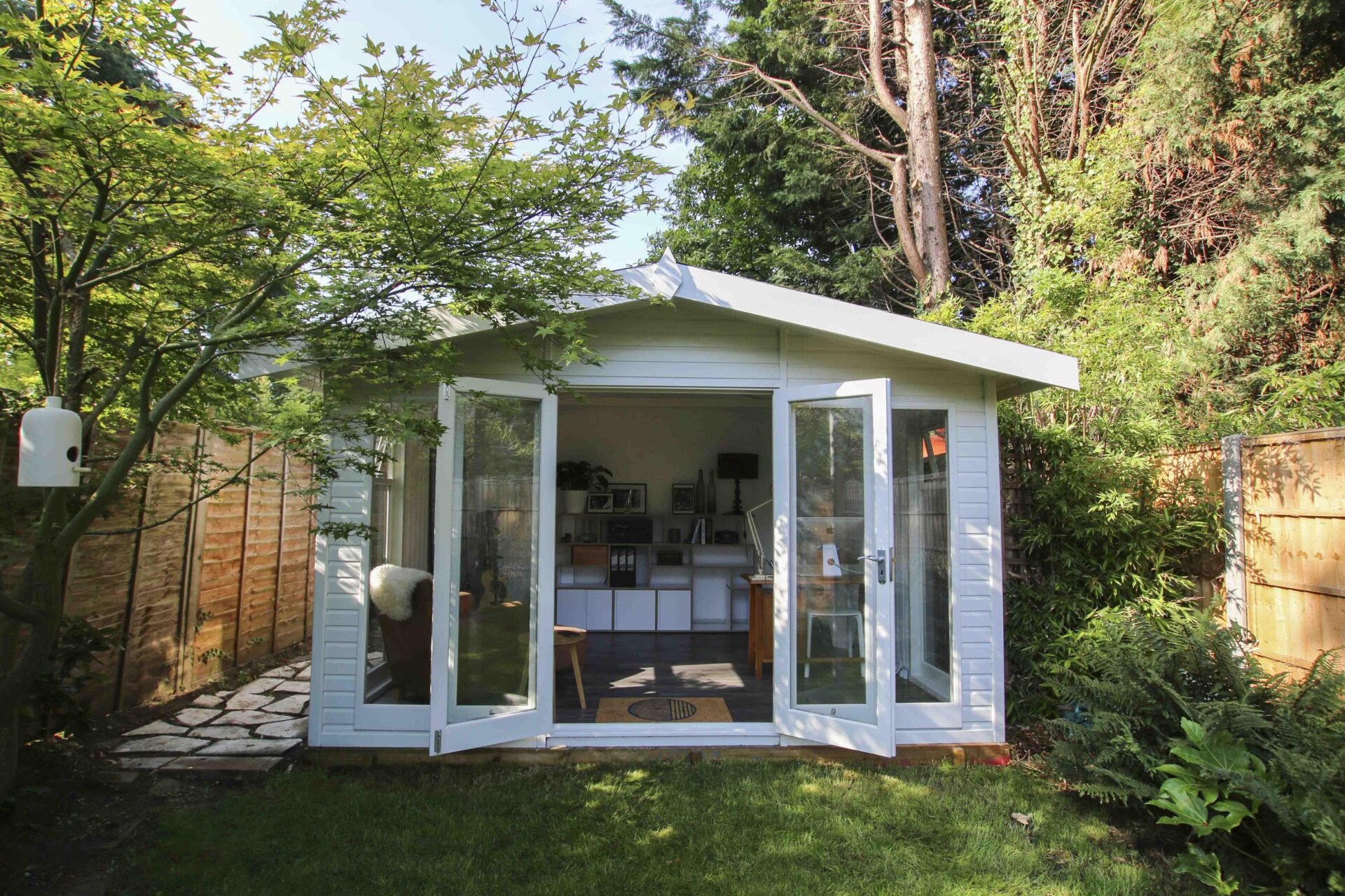 The image depicts a compact Studio Apex garden room by Malvern Garden Buildings, in a garden with a timber boundary fences. This customer used their garden building as a music room.