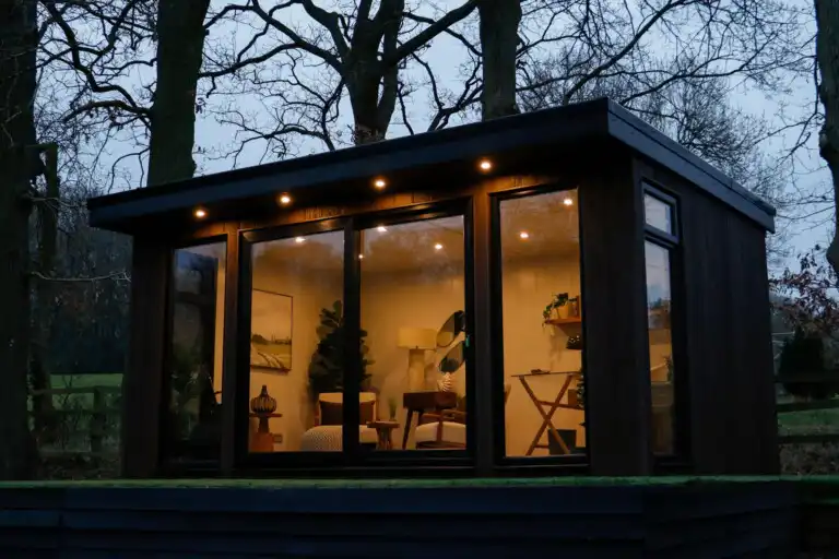 Malvern Garden Buildings composite garden room in wooden garden area, lit with exterior and interior lighting
