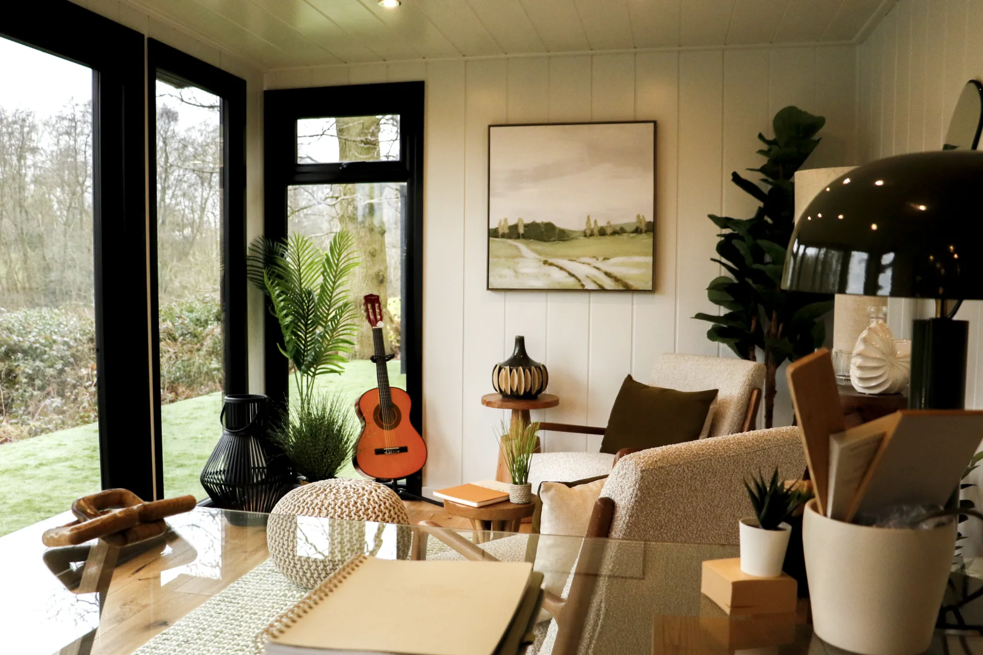 The image depicts the interior of an insulated garden office by Malvern Garden Buildings, featuring a desk and chairs, with large windows that provide a view of the garden.